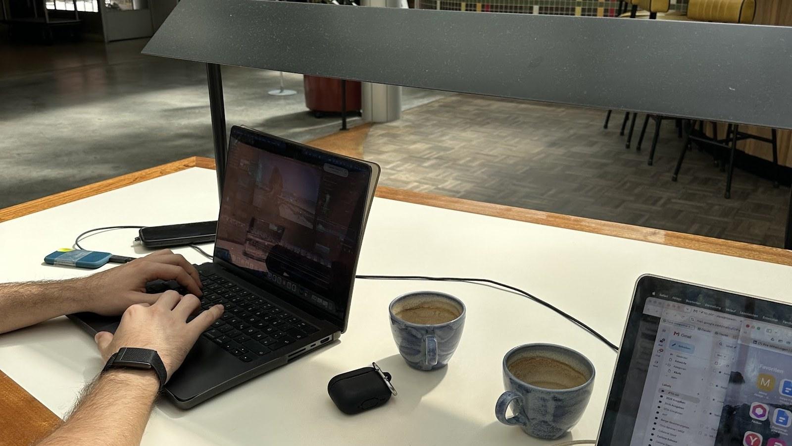 Zwei Laptops auf Holztisch im Coworking-Space, Hand am Trackpad